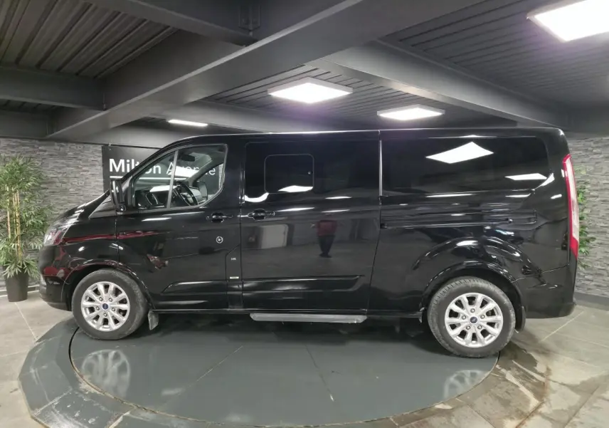 Vue de profil côté gauche d’un Ford Transit Fourgon Custom noir métallisé avec jantes alliage 17 pouces en intérieur showroom.