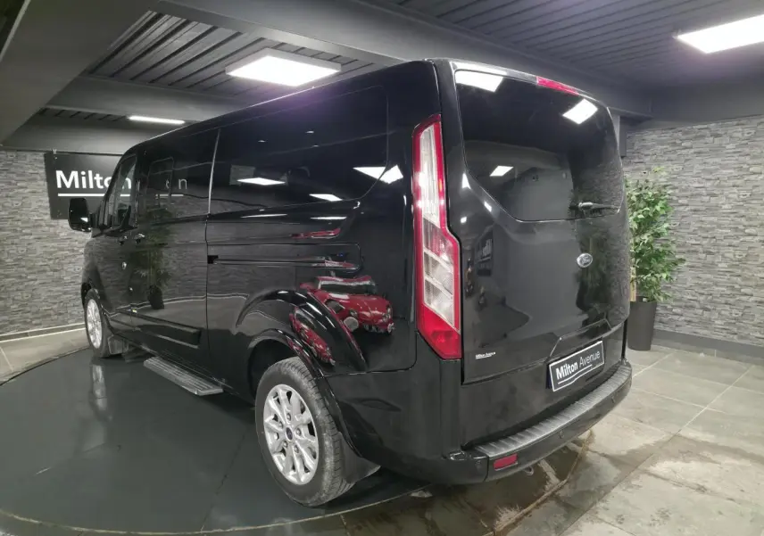 Vue 3/4 arrière droite du Ford Transit Fourgon Custom noir, avec jantes alliage et hayon vitré en intérieur showroom.
