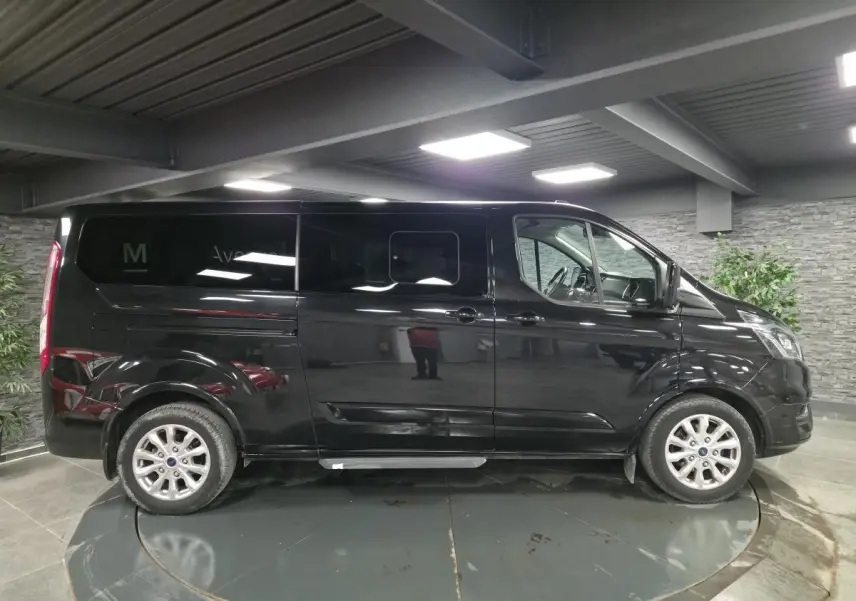 Vue de profil côté gauche d’un Ford Transit Fourgon Custom noir métallisé, avec jantes alliage et portes coulissantes.