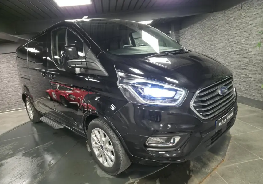 Ford Transit Fourgon Custom noir métallisé vu en 3/4 avant droit avec phares xénon allumés dans un garage.
