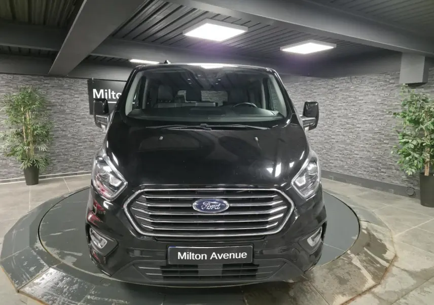 Vue frontale du Ford Transit Fourgon Custom noir, avec calandre chromée et phares allumés en showroom.