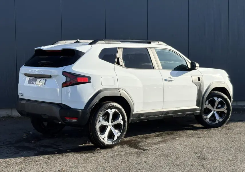 Vue 3/4 arrière droite du Dacia Duster blanc Glacier 2025 avec jantes alliage diamantées et barres de toit noires.