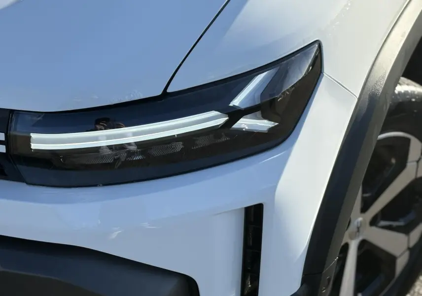 Gros plan sur le phare avant droit et la jante alliage diamantée d’un Dacia Duster blanc glacier 2025.