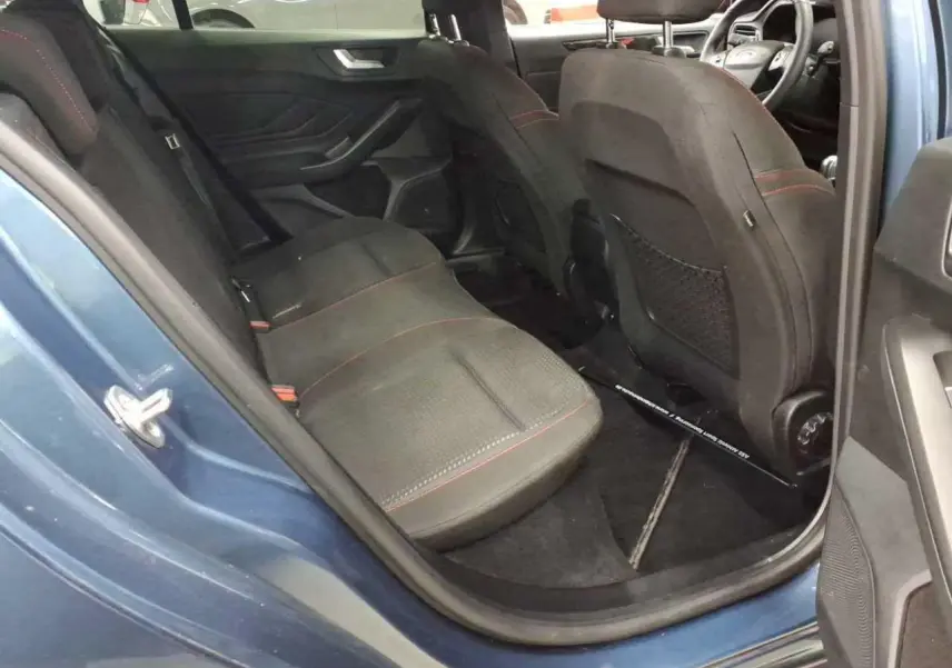 Vue intérieure côté arrière gauche de la Ford Focus SW Bleu Azur, sièges noirs avec surpiqûres rouges et volant visible.