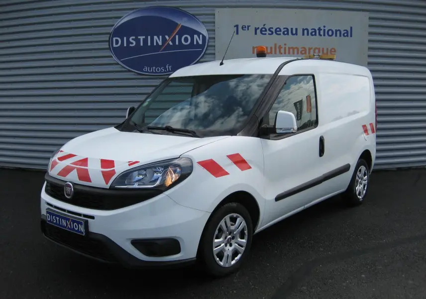 Vue 3/4 avant droit d’un FIAT Doblo Cargo blanc 2018 avec bandes rouges de signalisation et gyrophare orange sur le toit.