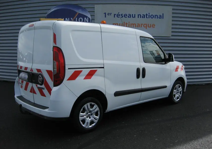 Fiat Doblo Cargo blanc vue 3/4 arrière droit avec bandes réfléchissantes rouges et blanches sur la carrosserie.