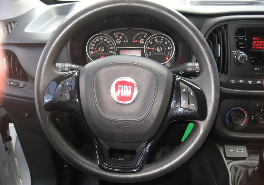 Vue rapprochée du volant noir avec logo FIAT rouge du Fiat Doblo Cargo blanc, tableau de bord et commandes visibles.
