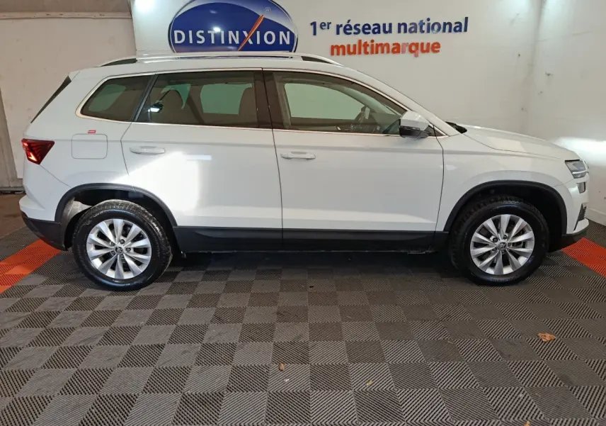 Profil gauche d'un SUV SKODA KAROQ blanc 2024, avec jantes alliage et barres de toit noires visibles en intérieur showroom.