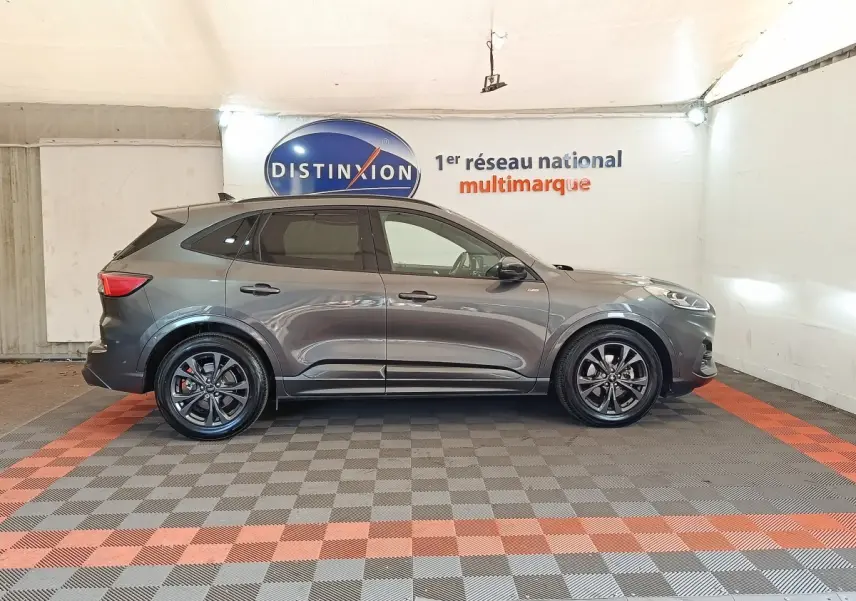 Ford Kuga hybride gris foncé vu de profil côté gauche, avec jantes noires et toit noir dans un showroom.