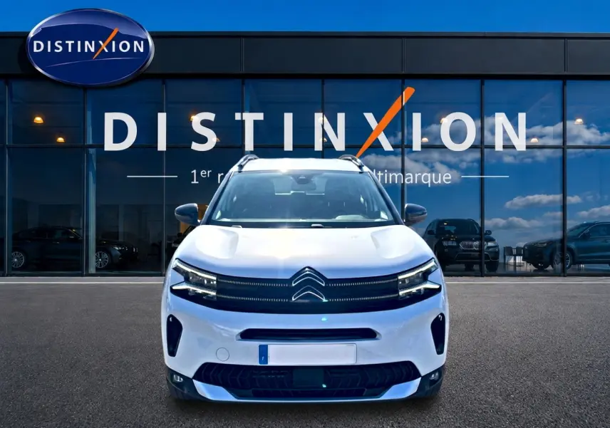 Vue avant d'un Citroën C5 Aircross blanc 2025 avec calandre distinctive et barres de toit visibles devant une vitrine.