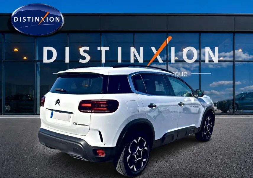 Vue 3/4 arrière droite d'un Citroën C5 Aircross blanc avec jantes alu et barres de toit devant un showroom.