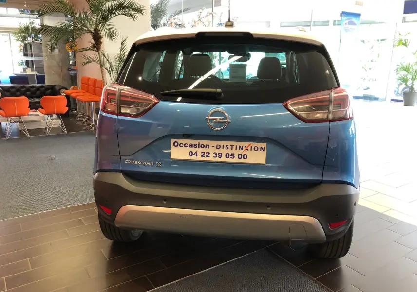 Vue arrière d'un Opel Crossland X bleu intens metal en showroom, avec logo et plaque d'occasion visibles.