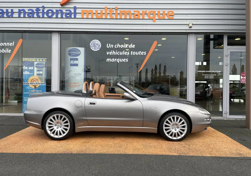 Profil droit d'une Maserati Spyder gris Nuvolari cabriolet de 2002 avec intérieur cuir beige, stationnée devant un showroom multimarque.
