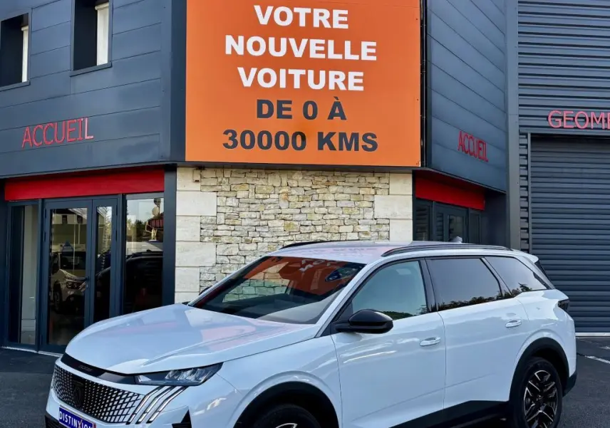 Peugeot 5008 blanc vue 3/4 avant droit, avec jantes noires et calandre distinctive, devant un bâtiment commercial.