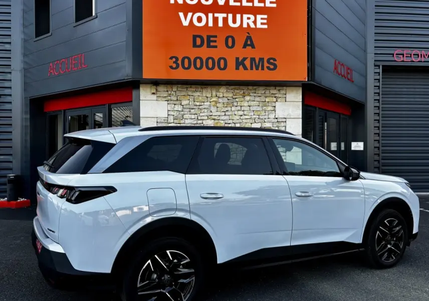Peugeot 5008 hybride blanc vue de profil droit, stationnée devant un bâtiment avec panneau orange "Votre nouvelle voiture".