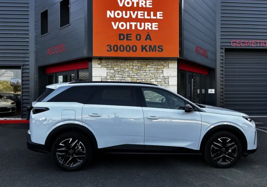 Profil droit du Peugeot 5008 blanc hybride 2025, avec jantes noires et vitres teintées, devant un bâtiment commercial.