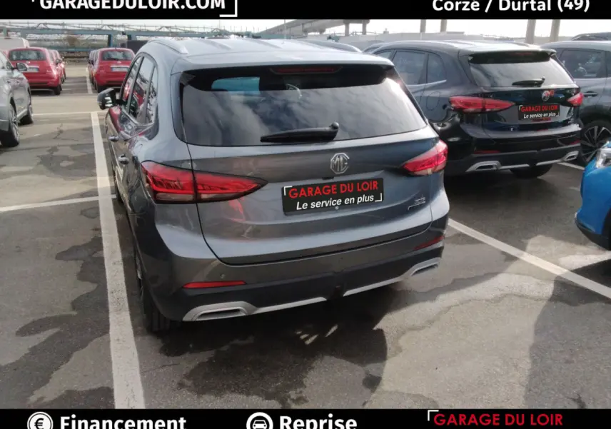 Vue 3/4 arrière d’un MG ZS gris clair hybride, avec feux arrière LED et logo MG visible sur un parking.