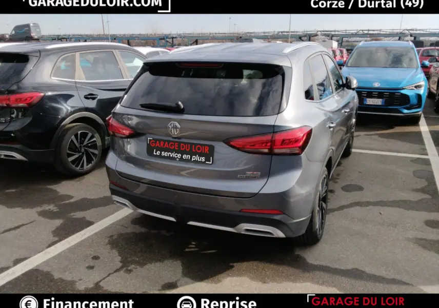 Vue 3/4 arrière d'un MG ZS gris clair hybride, avec feux arrière LED et logo MG visible sur un parking.