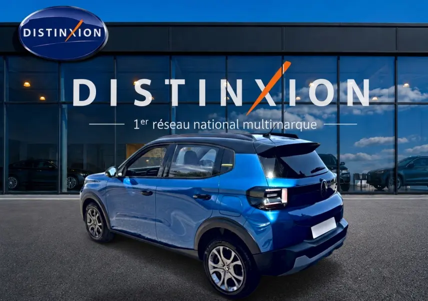 Vue 3/4 arrière droite d'une Citroën C3 2025 bleu avec toit noir et jantes alliage devant une vitrine Distinxion.