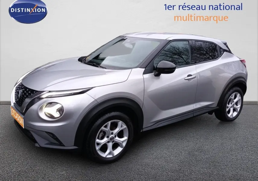 Nissan Juke gris platine métal en vue 3/4 avant droit, avec jantes alliage et phares allumés.
