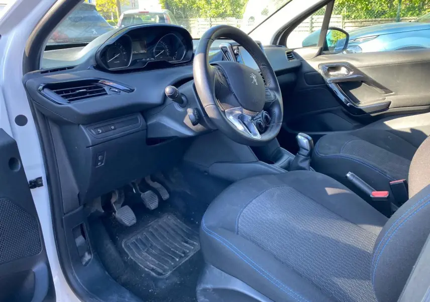 Vue intérieure côté conducteur de la Peugeot 208 blanche 2019, volant compact et sièges tissu avec surpiqûres bleues.
