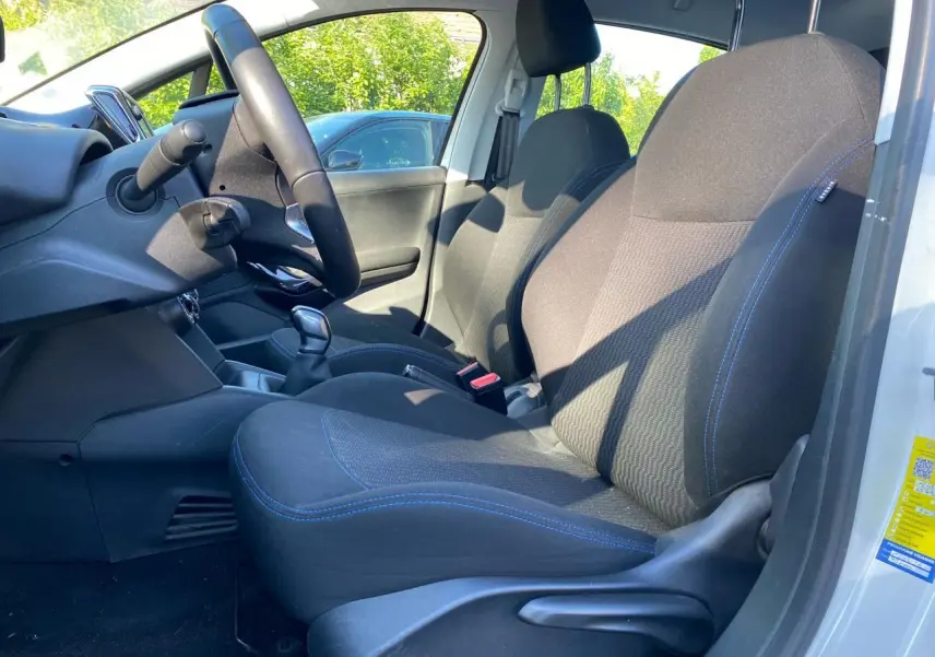 Vue intérieure côté conducteur de la Peugeot 208 blanche, montrant les sièges noirs avec surpiqûres bleues et le volant compact.