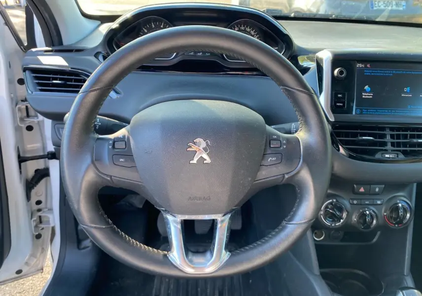 Vue intérieure centrée sur le volant Peugeot noir du 208 blanc, avec tableau de bord et écran tactile visible à droite.