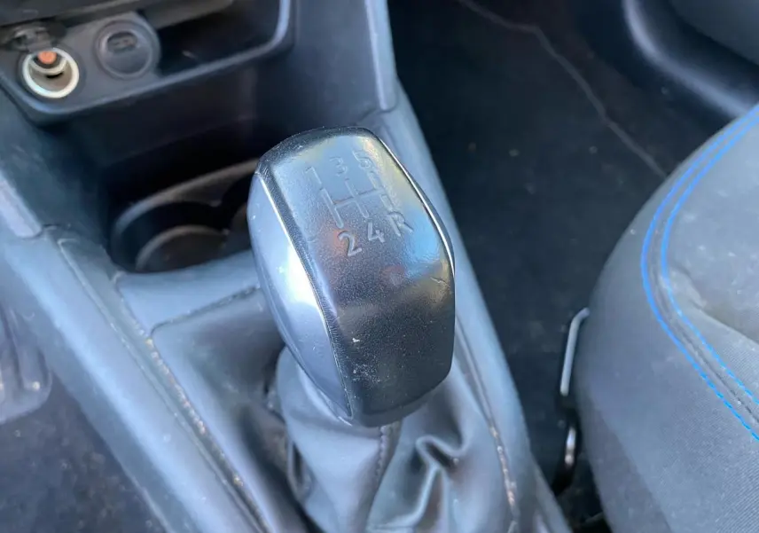 Gros plan sur le levier de vitesses manuel noir de la Peugeot 208 blanche, avec siège tissu gris et surpiqûres bleues visible à droite.