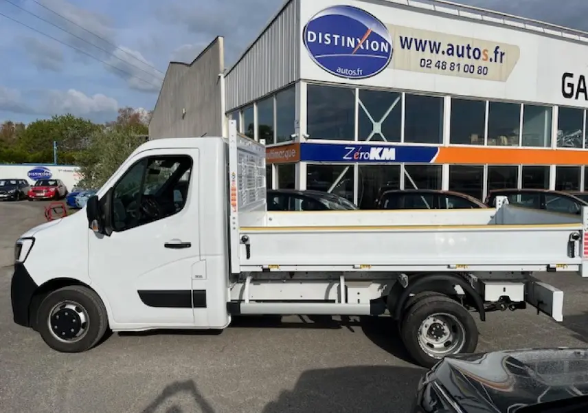 Vue latérale gauche d’un Renault Master blanc 2024 avec benne basculante, stationné devant un bâtiment commercial.