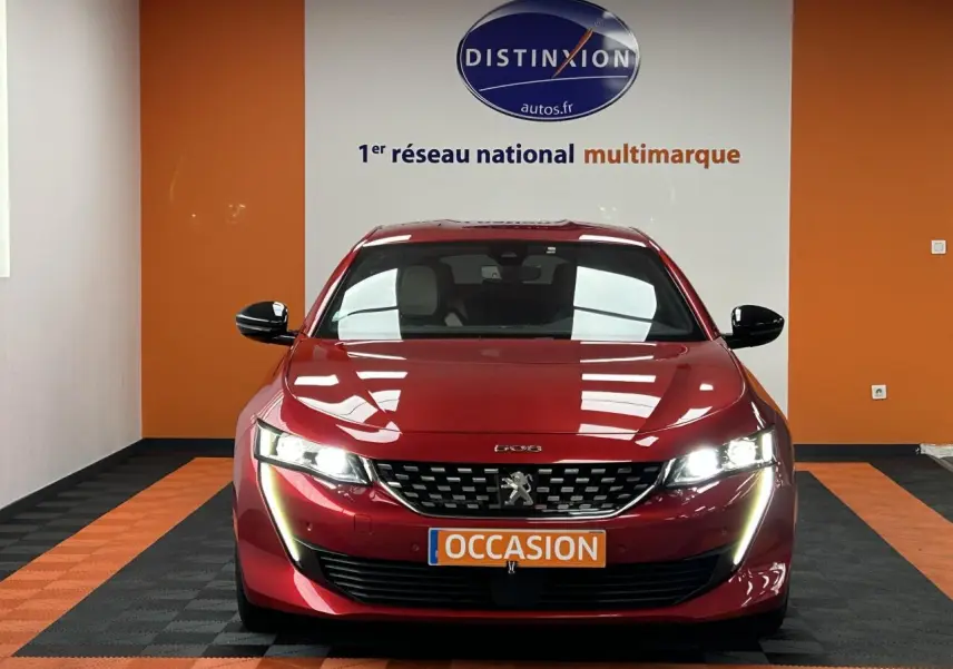 Vue frontale d'une Peugeot 508 Hybrid rouge clair avec phares allumés et plaque occasion dans un showroom.