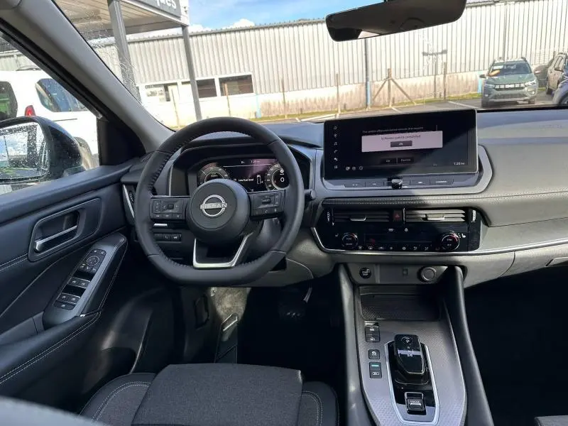 Vue intérieure avant du Nissan Qashqai Hybrid e-POWER 2025 en Gris Argile, avec tableau de bord et écran tactile central.