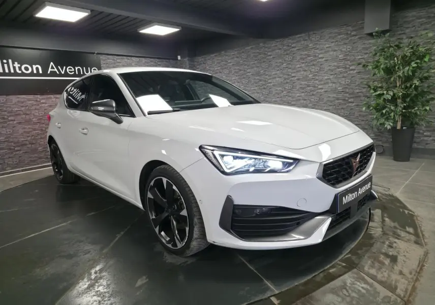 Vue 3/4 avant droite d'une CUPRA Leon blanche Nevada avec jantes noires et feux LED allumés en showroom.