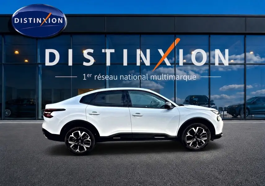 Profil droit d'une Citroën C4 X blanche 2025 avec jantes alu, devant une vitrine aux lettres DISTINXION.