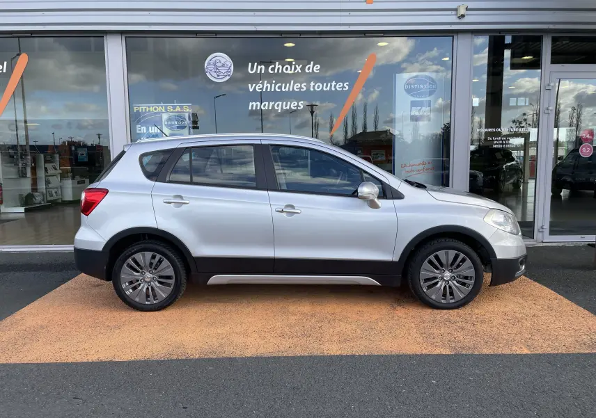 Profil côté gauche d'un Suzuki SX4 S-CROSS gris métallisé 2016 garé devant une vitrine de concession.