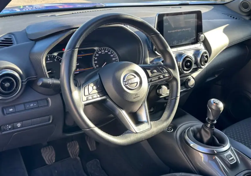 Vue intérieure du poste de conduite du Nissan Juke 2023.5 avec volant, écran tactile et levier de vitesses manuel.