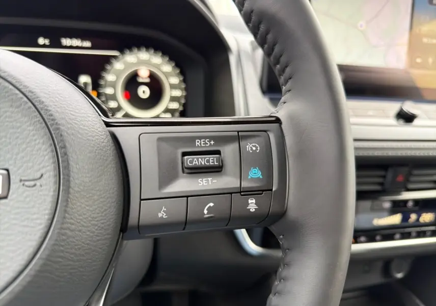 Gros plan sur les commandes au volant cuir noir du Nissan Qashqai 1.5 e-Power 2025, avec tableau de bord numérique en arrière-plan.