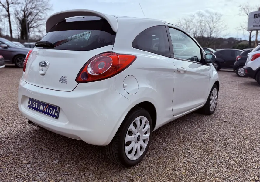 Vue 3/4 arrière droite d'une Ford Ka blanche avec hayon et jantes tôles, stationnée sur un sol gravillonné.