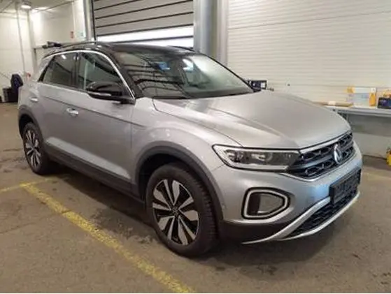 Volkswagen T-Roc gris clair en 3/4 avant droit, stationné en intérieur avec jantes distinctives et calandre moderne.