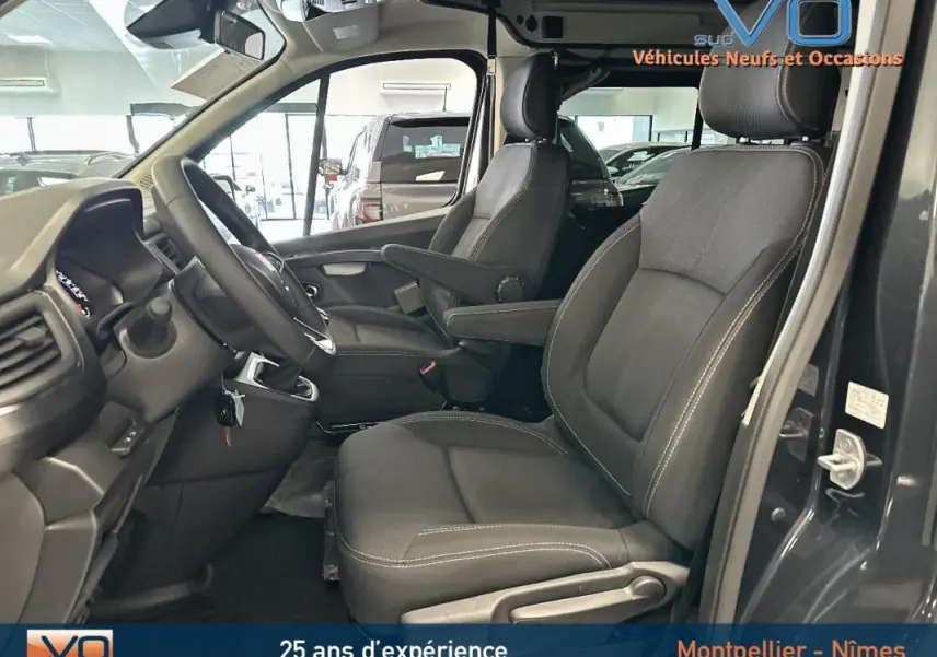 Intérieur côté conducteur du Renault Trafic Fourgon gris 2025, sièges tissu noirs avec accoudoirs et tableau de bord visible.
