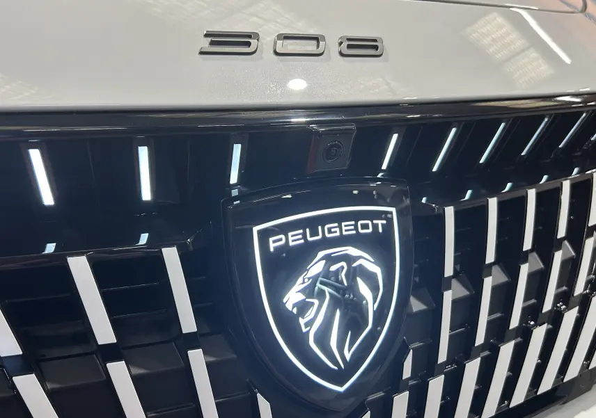 Gros plan sur la calandre noire et le logo lumineux Peugeot d’une 308 blanche Okenite, avec l’inscription 308 sur le capot.
