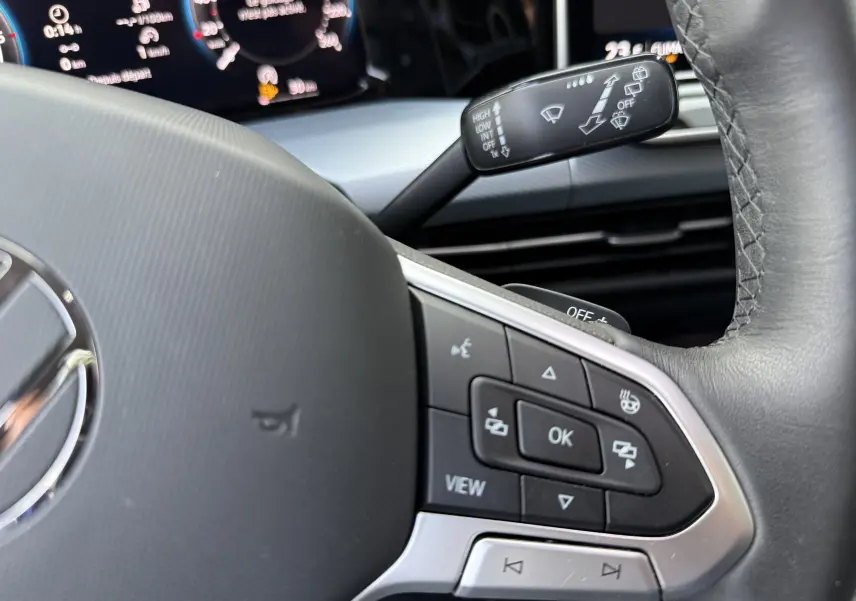 Gros plan sur le volant côté gauche d’une Volkswagen Golf SW gris lunaire, avec commandes multifonctions et tableau de bord numérique.
