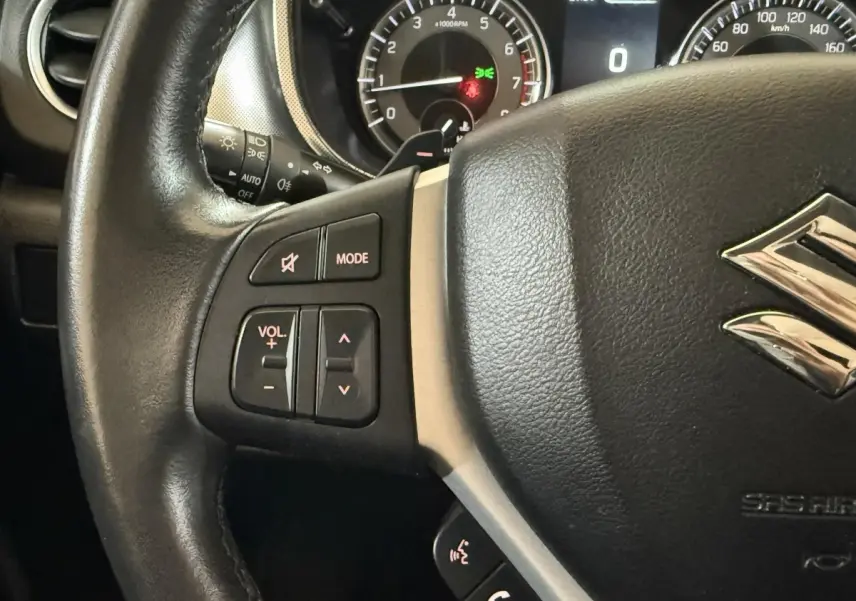 Gros plan sur le volant cuir noir du Suzuki Vitara 2022 avec commandes audio et tableau de bord éclairé en arrière-plan.