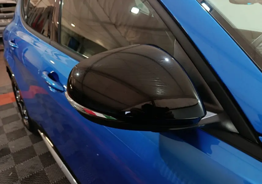 Gros plan sur le rétroviseur noir côté gauche d'une Alfa Romeo Tonale bleu 2023, avec reflet du plafond visible.