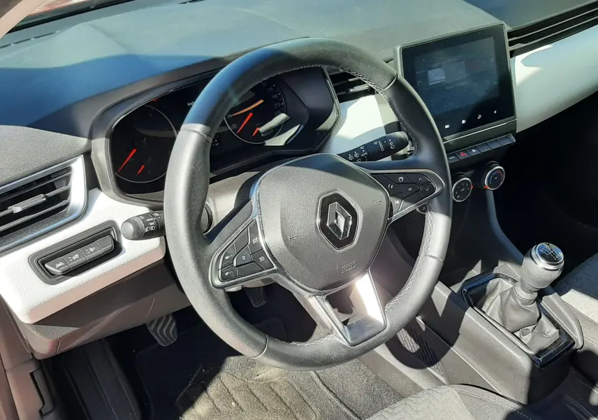 Vue intérieure de face du poste de conduite d'une Renault Clio V rouge, volant multifonction et boîte manuelle visibles.