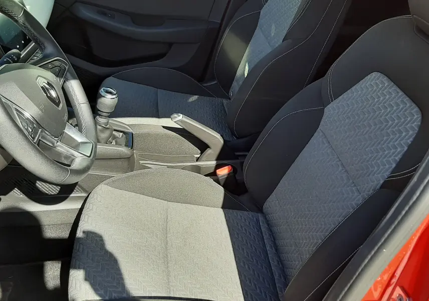 Vue intérieure côté conducteur de la Renault Clio V rouge, montrant les sièges tissu gris noir et la boîte manuelle.