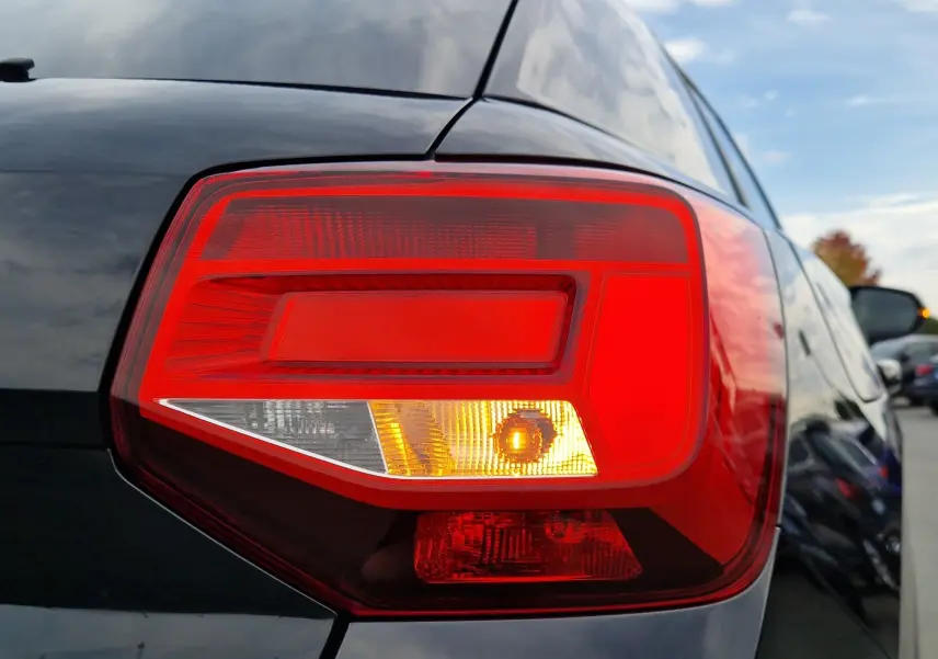 Gros plan sur le feu arrière droit rouge et orange d’un Audi Q2 noir mito metal 2023, avec reflet du ciel et voitures alentour.