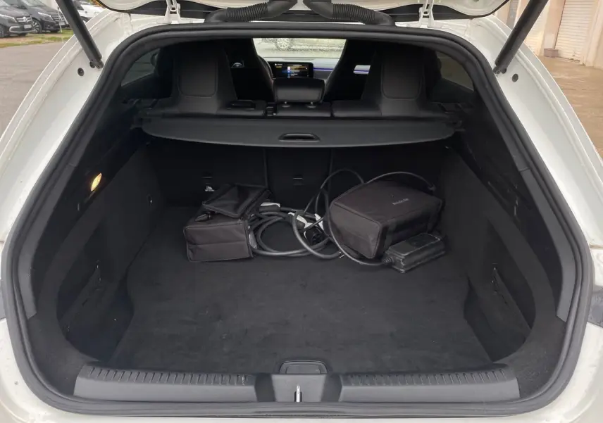 Coffre ouvert d'une Mercedes CLA Shooting Brake blanche, vue arrière montrant l'espace de chargement et câbles de recharge hybride.
