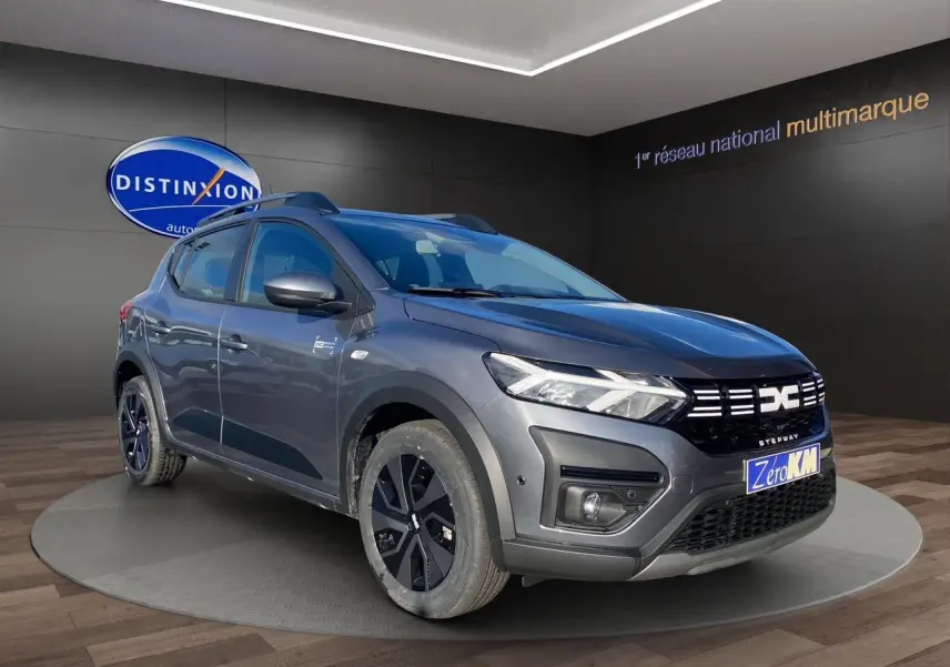 Dacia Sandero Stepway gris schiste en 3/4 avant droit, avec barres de toit et jantes noires distinctives.