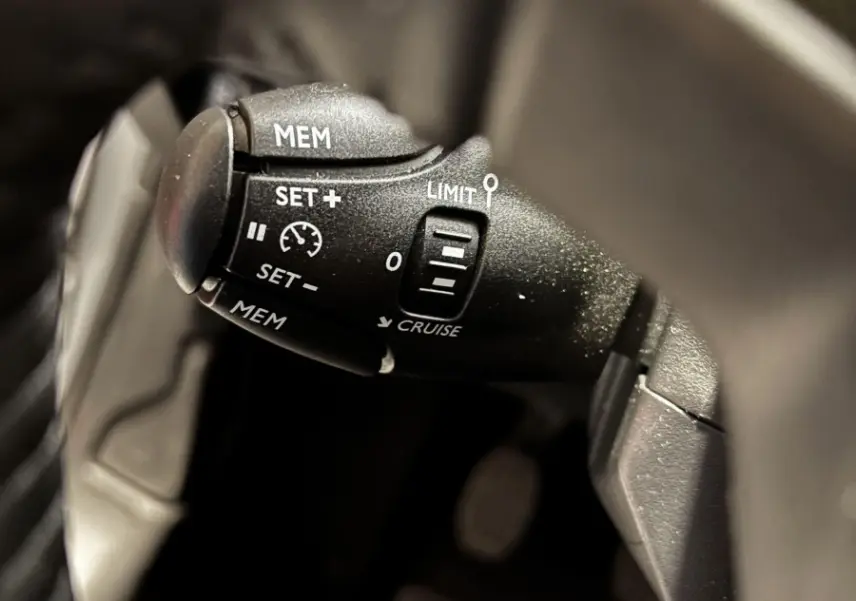 Gros plan sur la commande de régulateur de vitesse noire à gauche du volant d'une Peugeot 208 gris clair 2024.