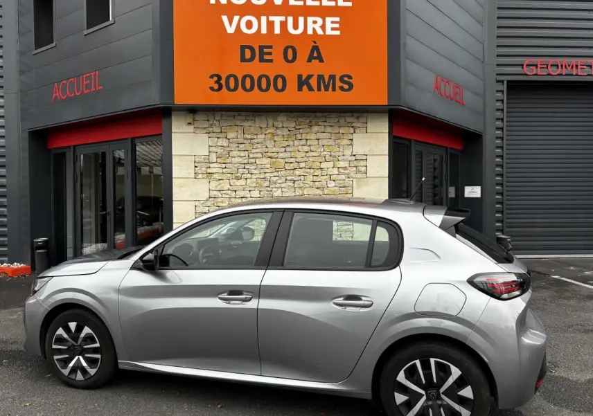 Peugeot 208 PureTech 100 S&S Active gris clair vue de profil côté gauche devant un bâtiment avec panneau orange.